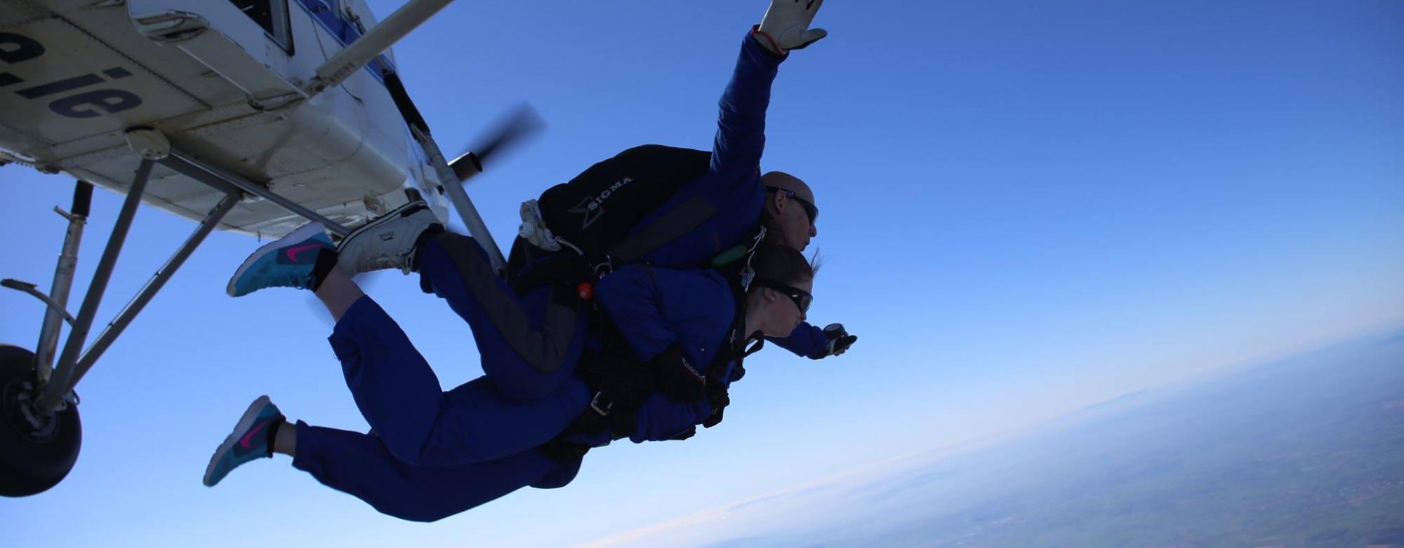 Blue sky with part of a plane visible, and two people wearing parachutes