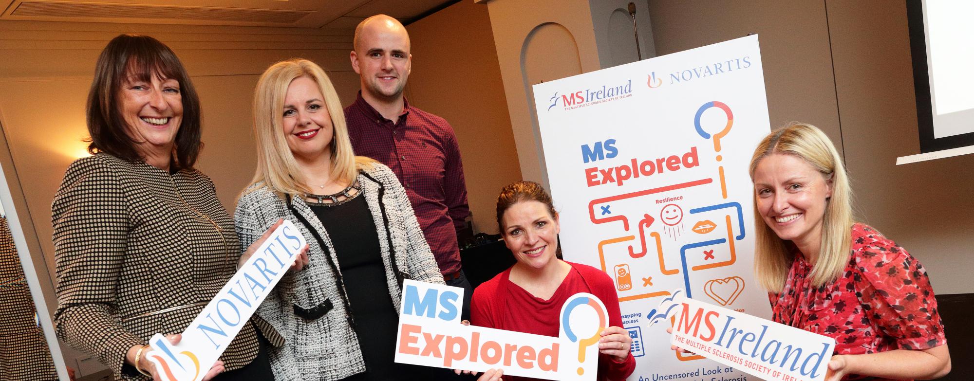Banner in the background with three individuals standing, two sitting in front, and two holding signage for MS Research Explored 2.