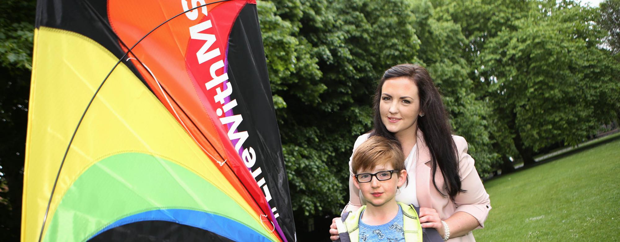 Photo of a mother with dark hair wearing a pink jacket and her son wearing black glasses, a blue t-shirt and dark jacket and holding a multicoloured kite in a park. t