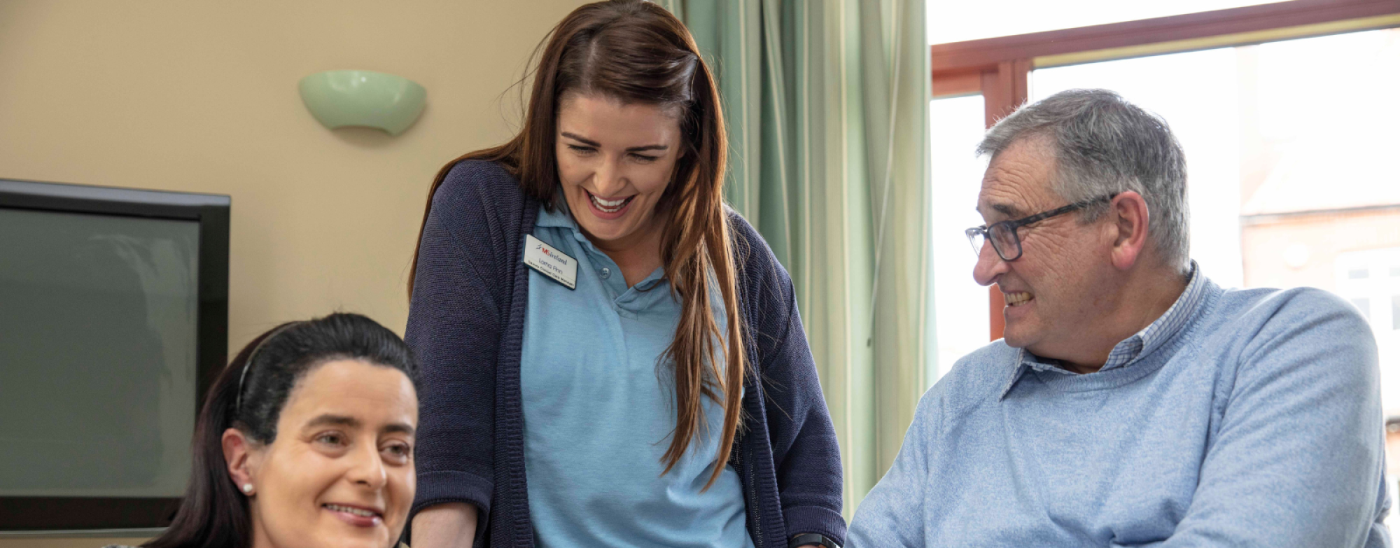 Two individuals are seated while one person stands. In the background, there is a television and a window