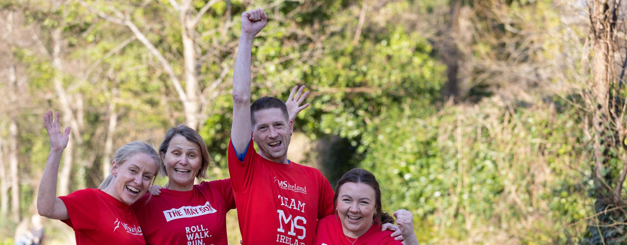 Four people in red “May 50K” shirts cheer on a tree-lined path.