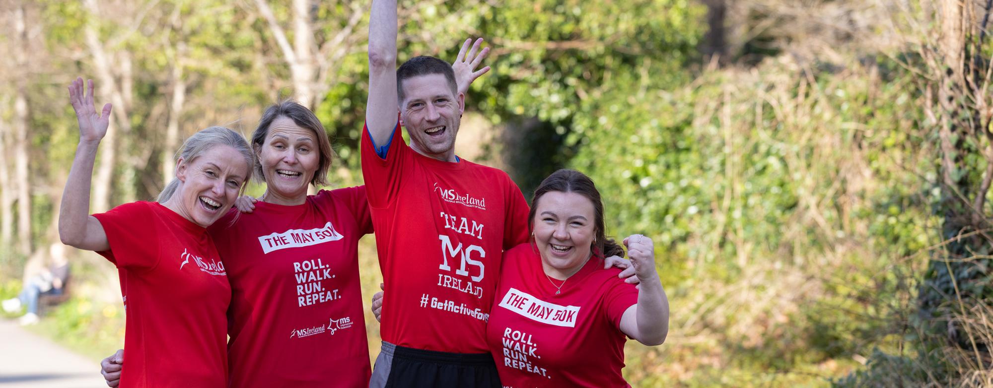 Four people in red “May 50K” shirts cheer on a tree-lined path.