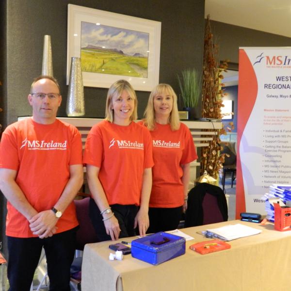 Image of three individuals, one male, two female standing behind a registration desk. All three are wearing red t-shirts with white MS Ireland logos. 