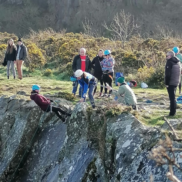 A group of people stands at the edge of a cliff, geared up with helmets, harnesses, and ropes, preparing to abseil down. The backdrop features a panoramic view of a rugged landscape, with a clear blue sky overhead