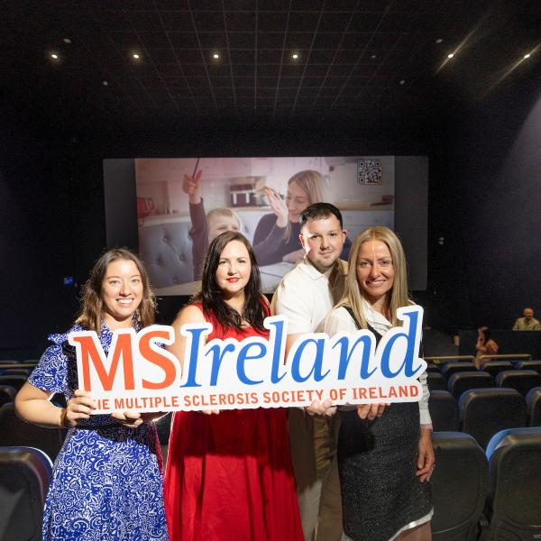 Four people holding MS Ireland signage in front of a cinema screen displaying a World MS Day awareness ad.