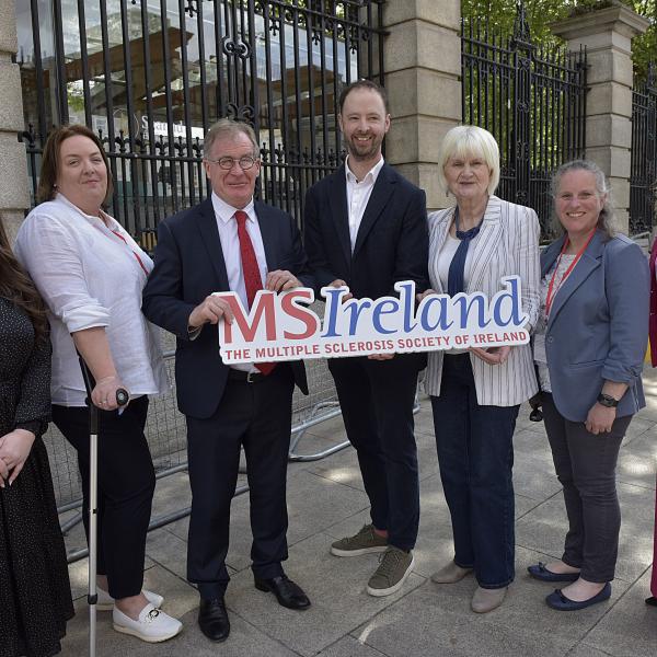 Seven people standing outside Leinster House, Dublin, holding an MS Ireland banner after a briefing with Oireachtas Members.