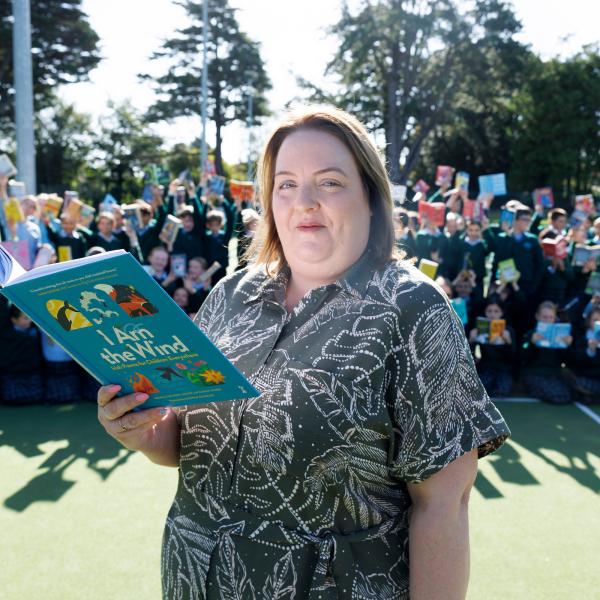 Louise Power, holding a children’s book titled I Am the Wind, stands outdoors with a large group of schoolchildren behind her raising their books to celebrate the MS Readathon