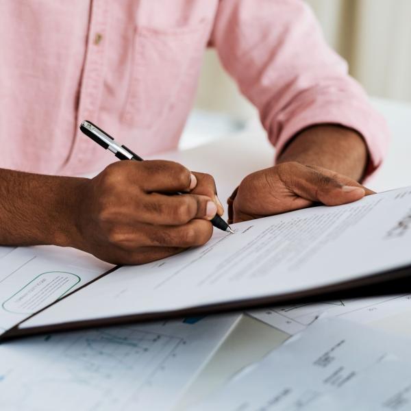 A person writing on a document with a pen on a clipboard, with papers spread on a desk.