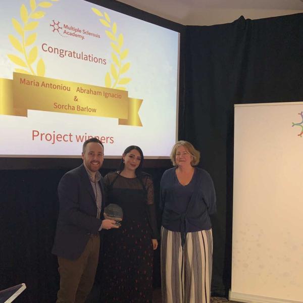 Three people standing on a stage holding an award in front of a presentation slide announcing project winners, with a Neurology Academy banner beside them