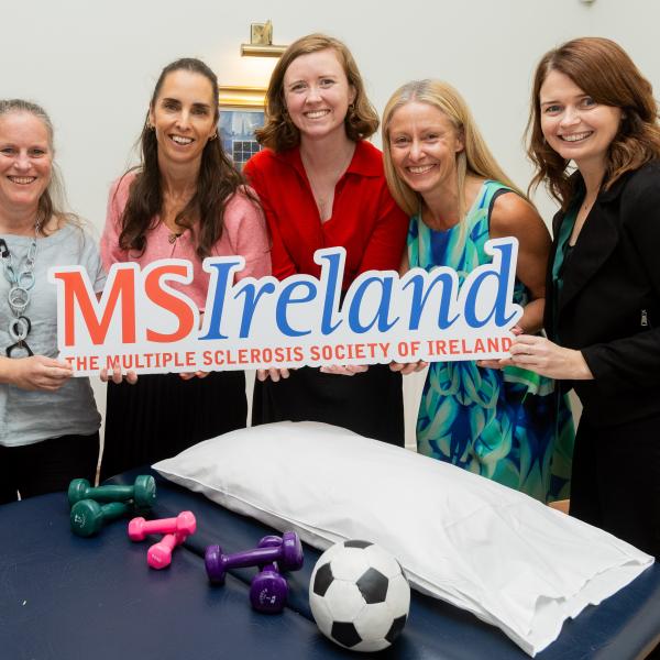Five people holding an MS Ireland sign beside physiotherapy equipment, including dumbbells, a ball and a treatment table.