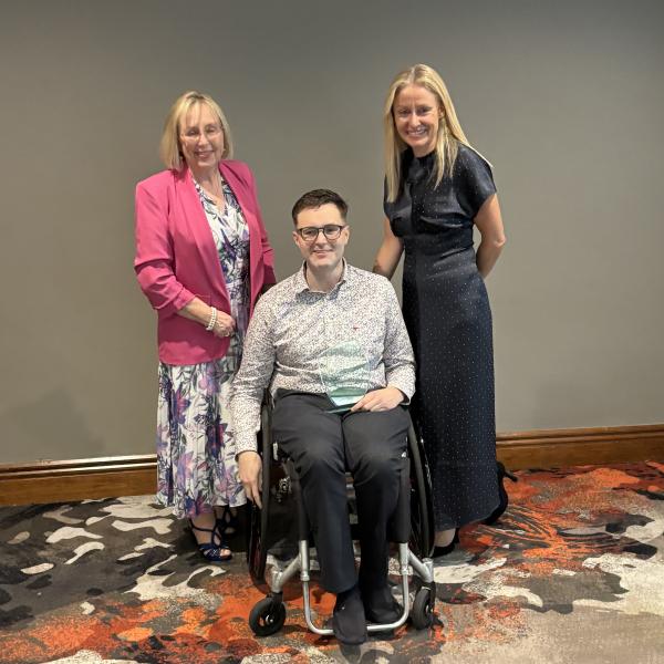 Billy, MS Ireland's 2026 Person of the Year, seated in his wheelchair holding his award, alongside two women at the awards ceremony.
