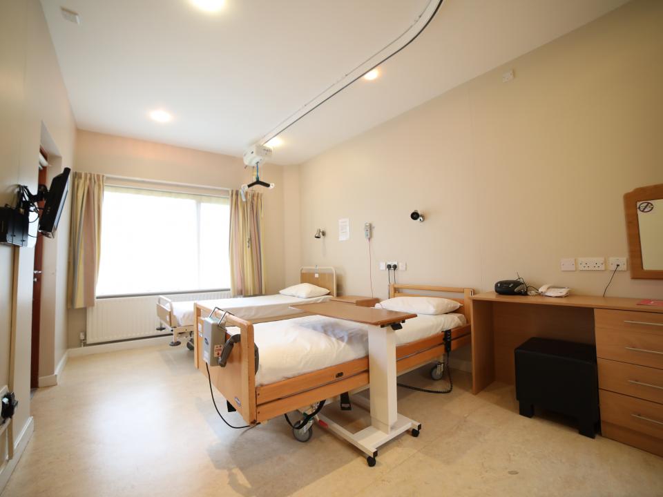 A bedroom in an MS care centre, featuring a bed with a hoist, a locker for storage, and a large window letting in natural light, creating a comfortable and accessible environment