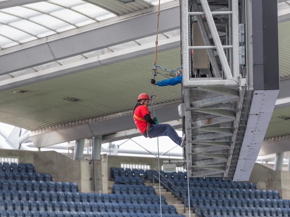 A person wearing a red t-shirt abseiling down from a great height at Croke Park, with a clear view of the stadium and ground far below