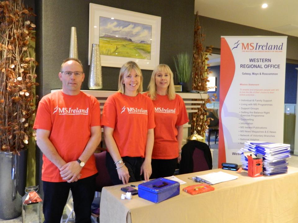 Image of three individuals, one male, two female standing behind a registration desk. All three are wearing red t-shirts with white MS Ireland logos. 