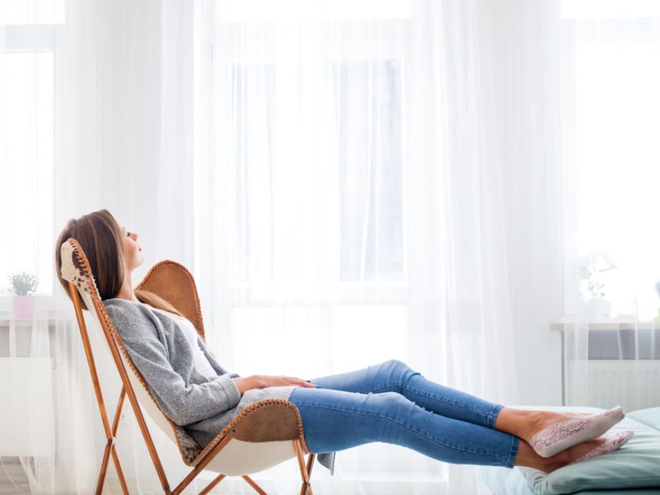 Image of a lady in blue jeans and a light top reclined on a chair with her feet resting on a stool with white curtains in the background.