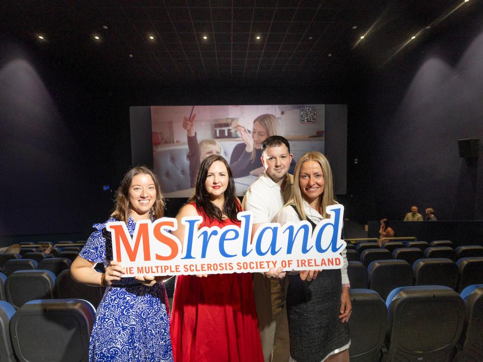Four people holding MS Ireland signage in front of a cinema screen displaying a World MS Day awareness ad.