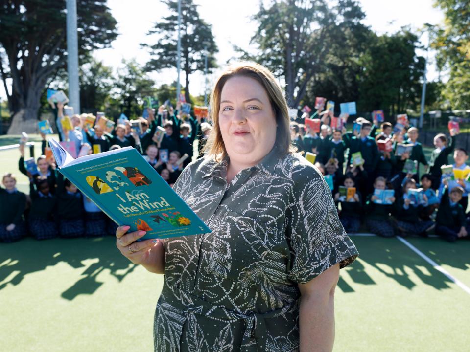 Louise Power, holding a children’s book titled I Am the Wind, stands outdoors with a large group of schoolchildren behind her raising their books to celebrate the MS Readathon