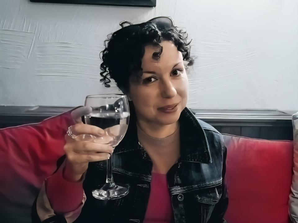 Woman with short dark curly hair raising a glass of water while seated at a table