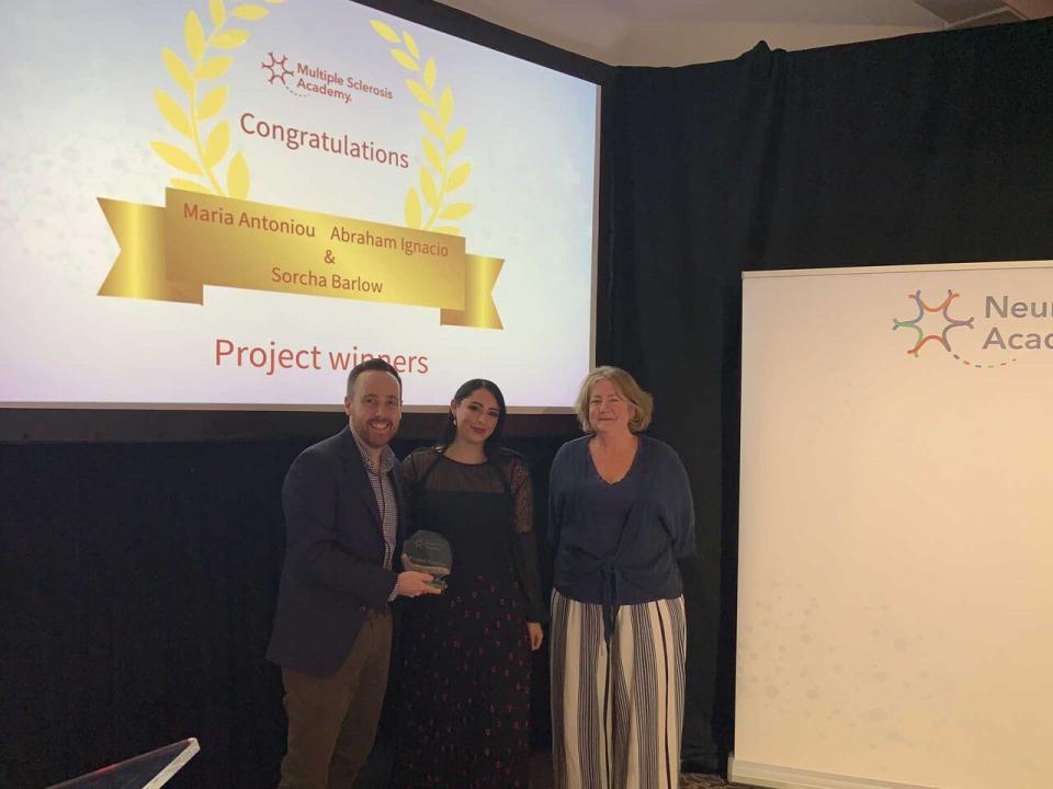 Three people standing on a stage holding an award in front of a presentation slide announcing project winners, with a Neurology Academy banner beside them