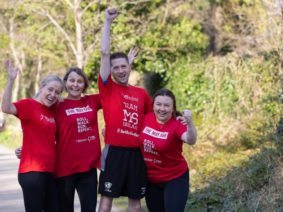 Four people in red “May 50K” shirts cheer on a tree-lined path.