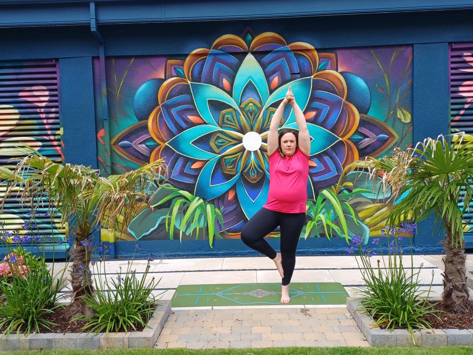 Áine Gegarty balancing in a yoga pose on a mat outdoors, with a vibrant mural in the background.