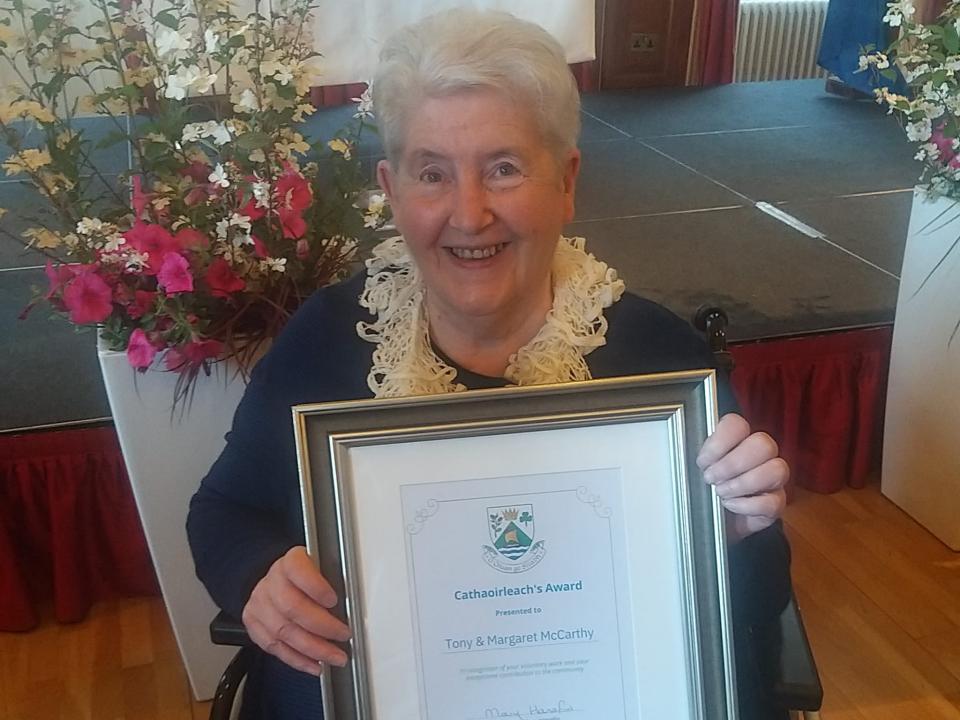 Margaret McCarthy smiling and holding a framed Cathaoirleach’s Award while seated in a wheelchair.