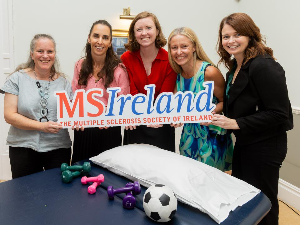 Five people holding an MS Ireland sign beside physiotherapy equipment, including dumbbells, a ball and a treatment table.