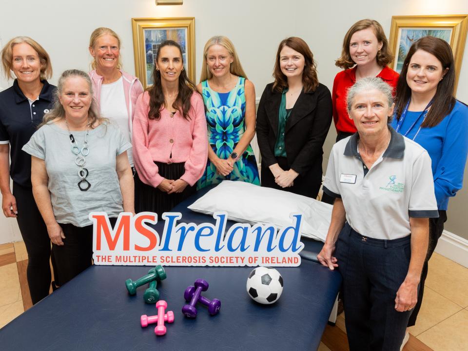Nine people gathered around a physiotherapy table with an MS Ireland sign, dumbbells and a ball.