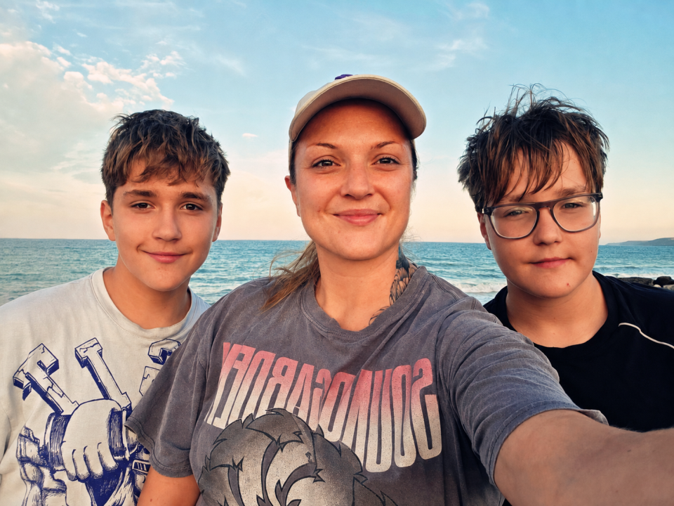 A woman wearing a cap takes a selfie with her two sons by the sea.