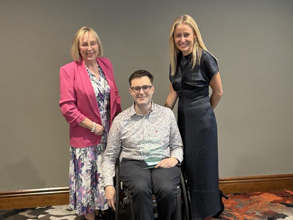 Billy, MS Ireland's 2026 Person of the Year, seated in his wheelchair holding his award, alongside two women at the awards ceremony.