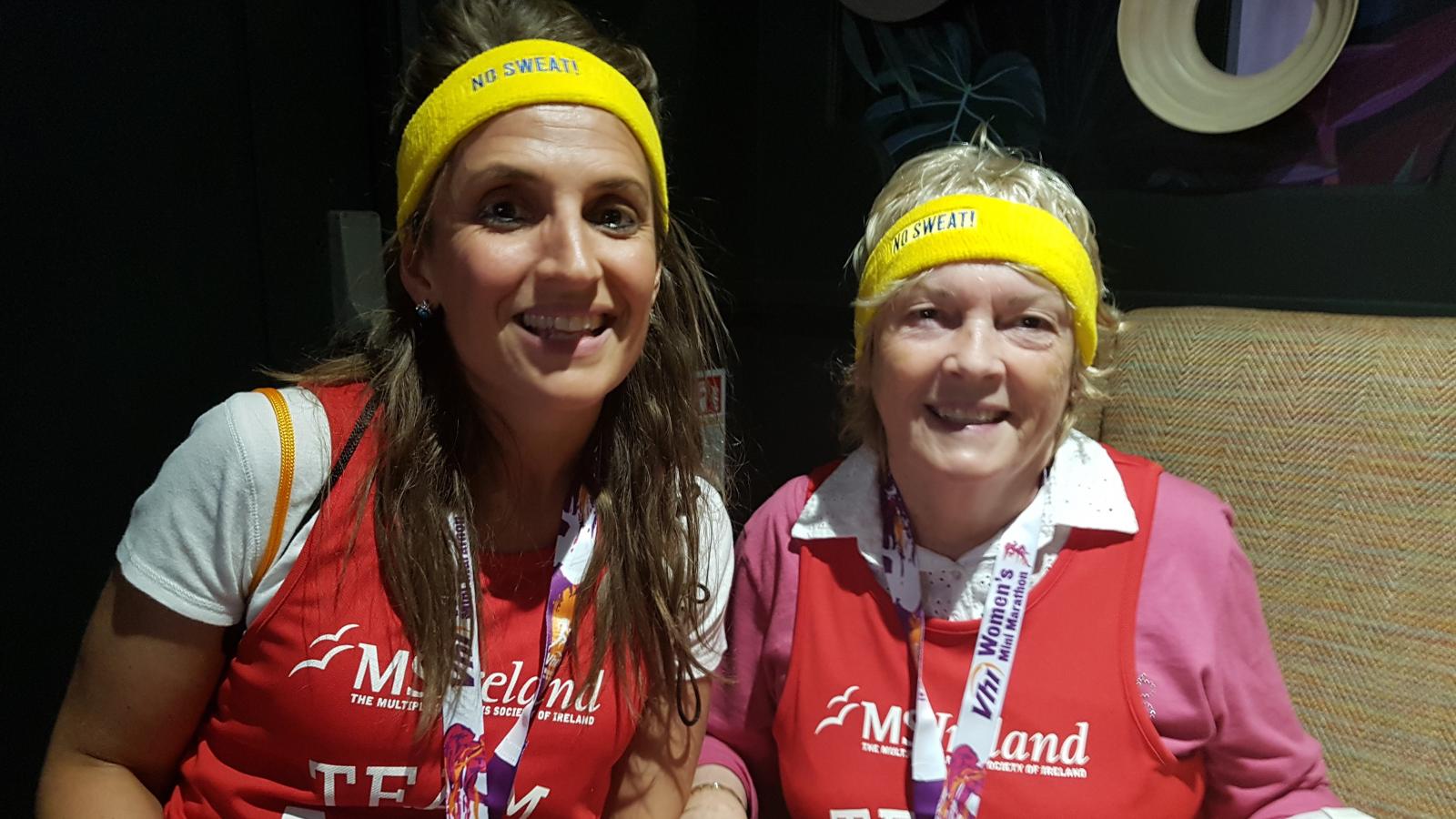 Two people smiling, wearing sweatbands and red MS Ireland t-shirts, with medals around their necks.
