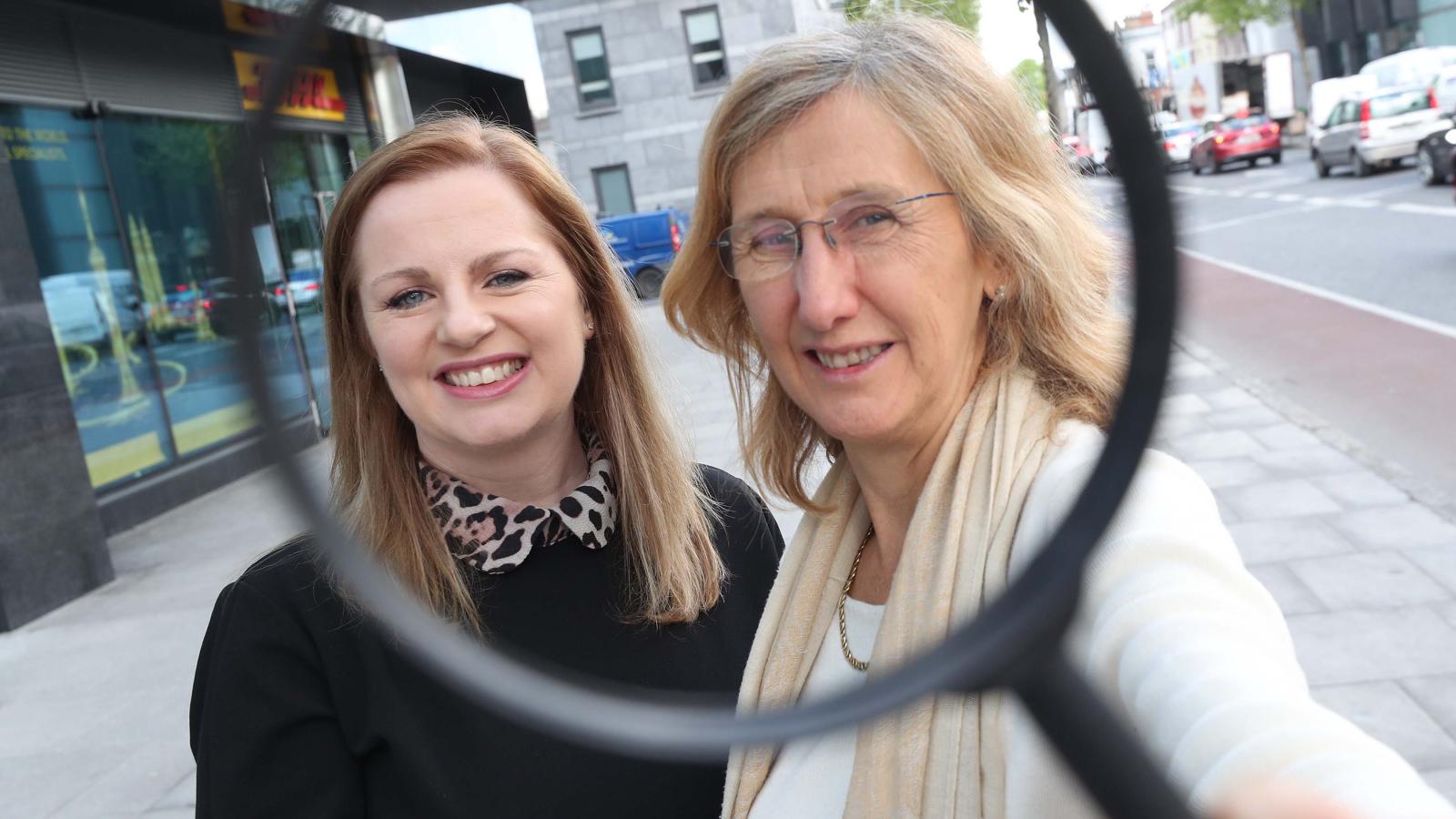Two people standing together, with one holding a large magnifying glass in front of the camera, symbolising research and investigation