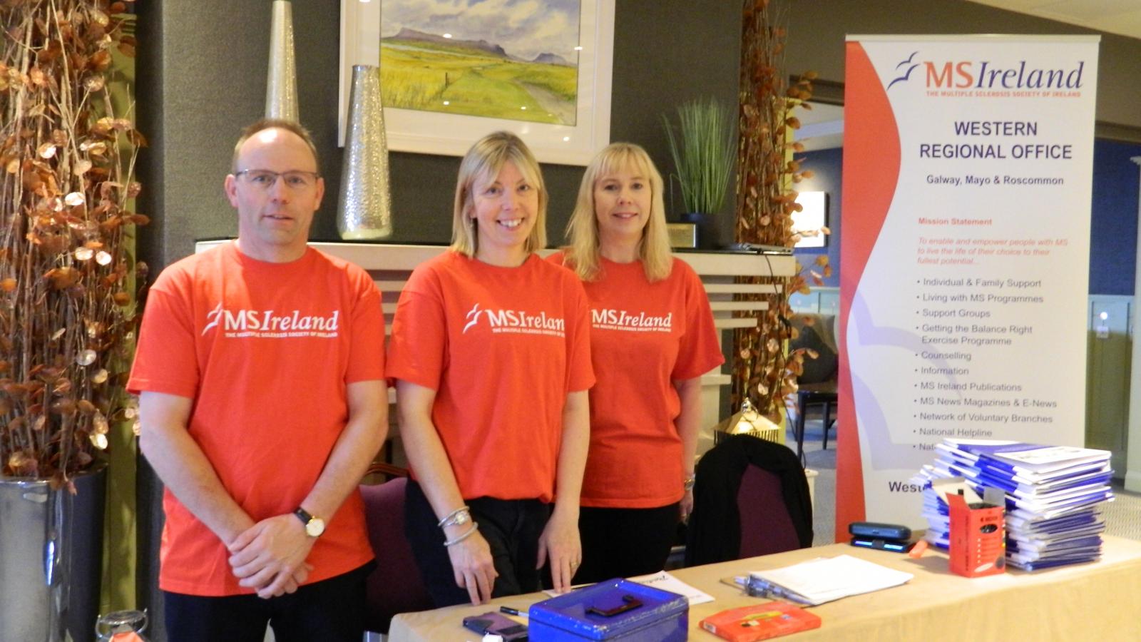 Image of three individuals, one male, two female standing behind a registration desk. All three are wearing red t-shirts with white MS Ireland logos. 