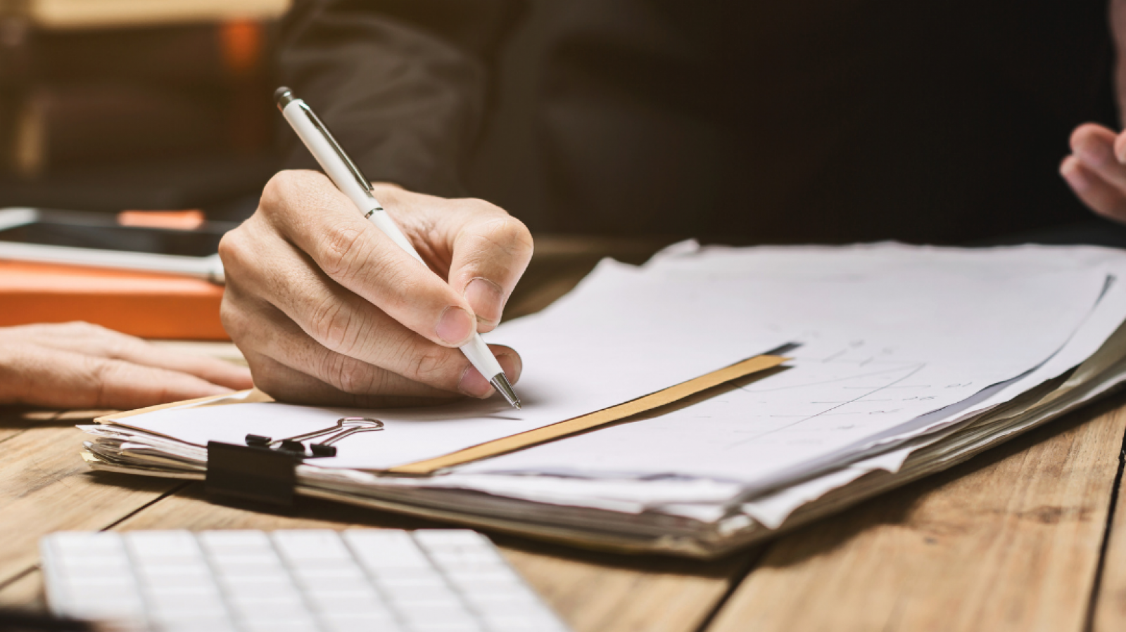 Someone with a dark top writing with a pen onto paper on a clipboard at a table.