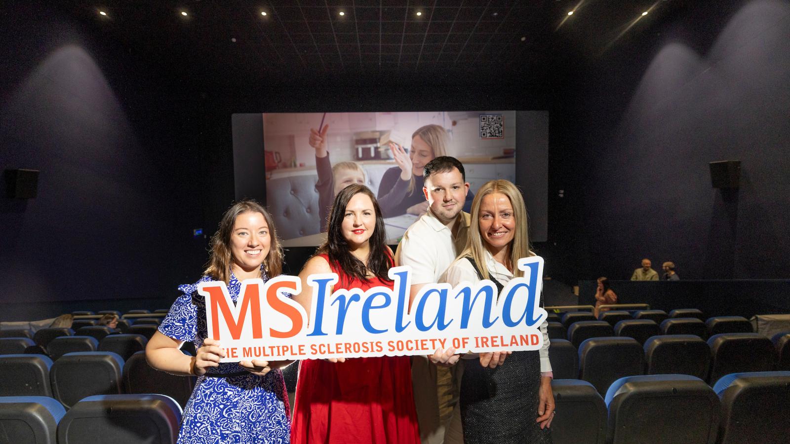 Four people holding MS Ireland signage in front of a cinema screen displaying a World MS Day awareness ad.