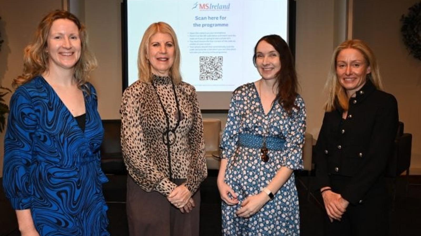Four people standing together at a PROMISE Project event, with an MS Ireland presentation slide displayed on a screen behind them.