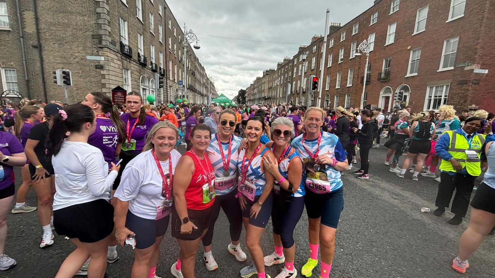 A group of women runners stand together on a city street after a race, smiling with medals around their necks as crowds fill the background.