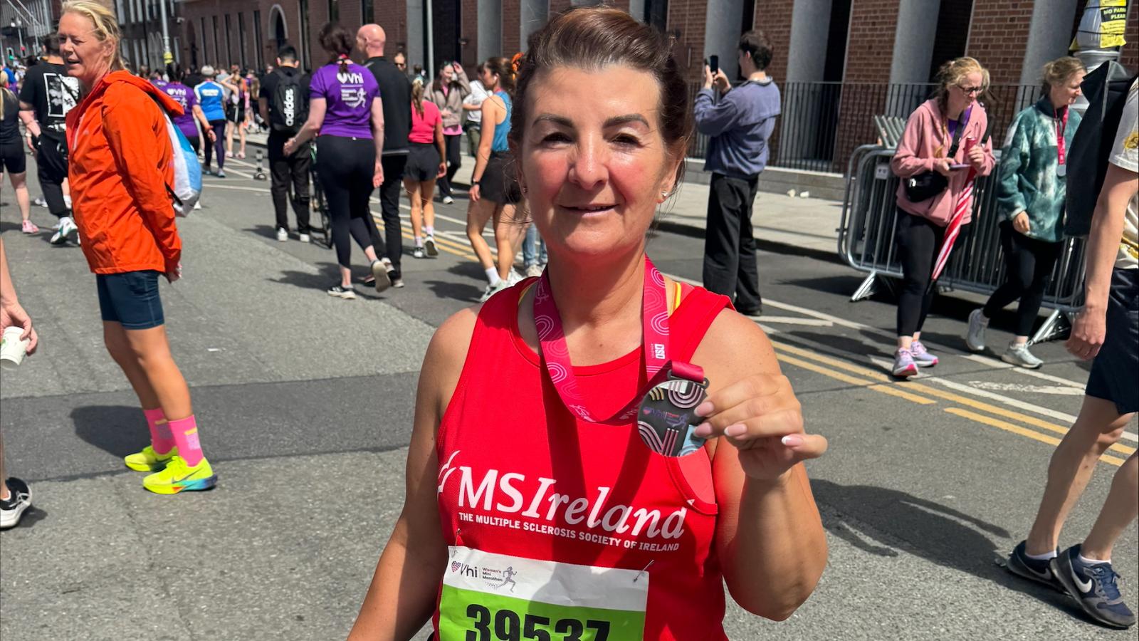 A smiling runner in a red MS Ireland vest holds up her finisher’s medal on a sunny city street, with other runners in the background.
