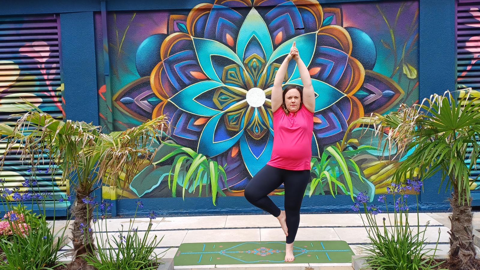 Áine Gegarty balancing in a yoga pose on a mat outdoors, with a vibrant mural in the background.